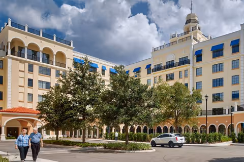 Exterior view of a large multi-story senior living facility with cream-colored walls and blue awnings. There are trees and landscaping in front of the building, a white car parked near the entrance, and two elderly people walking on the sidewalk. The sky is partly cloudy.