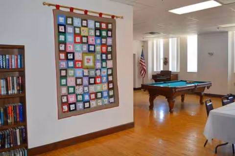 Interior lounge with quilt wall hanging, bookshelf, pool table, and American flag in a senior facility.