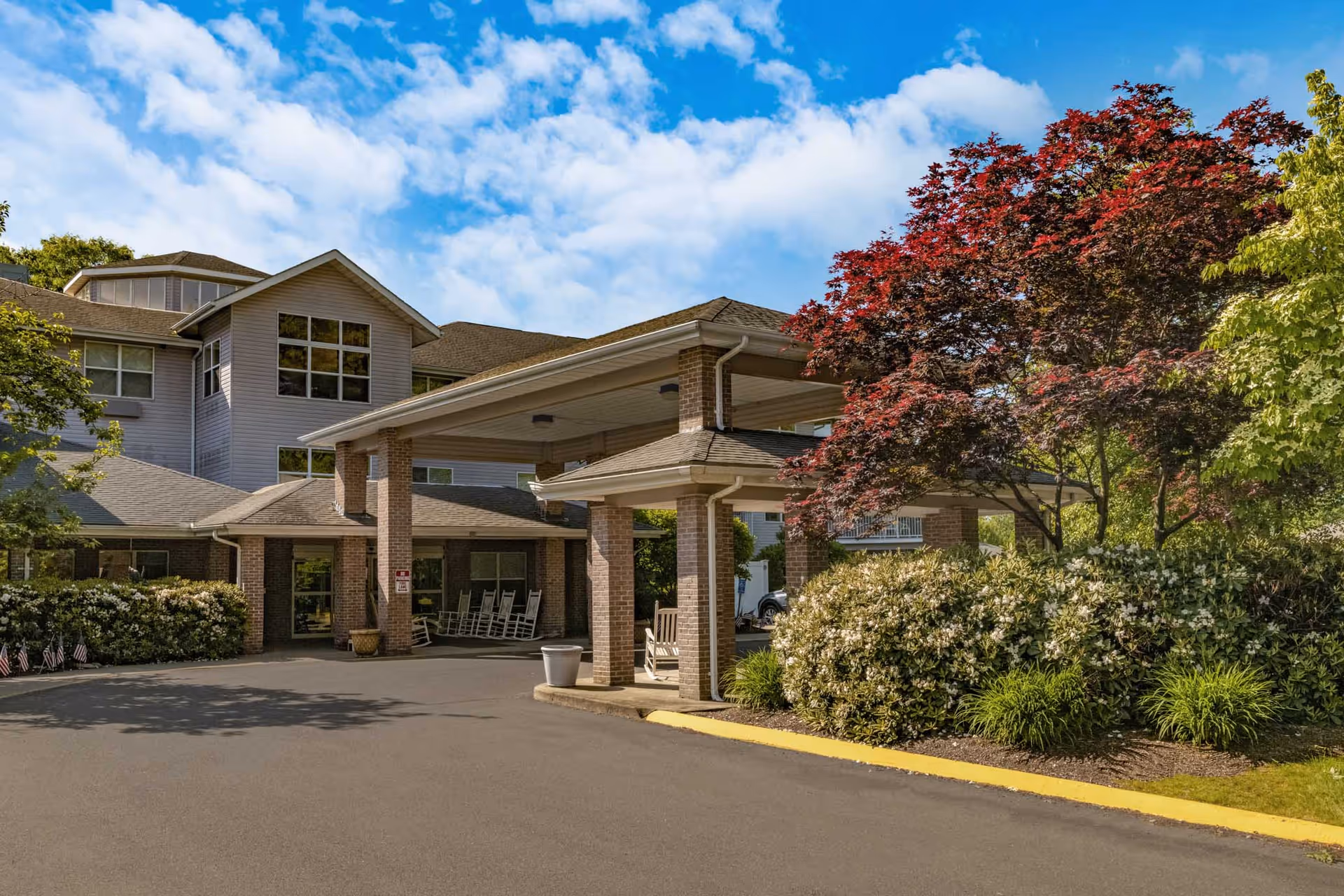 Exterior view of Holiday Quail Run Estates showing the entrance with a covered driveway supported by brick columns, surrounded by well-maintained bushes and trees under a partly cloudy sky.