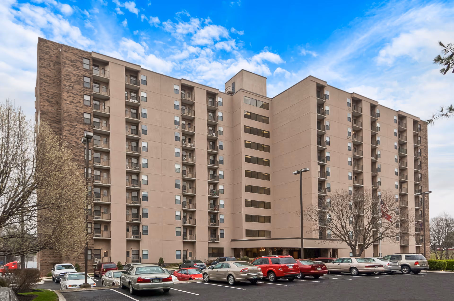 Exterior front view of a multi-story senior living apartment building with balconies and a parking lot of cars in front under a partly cloudy sky.