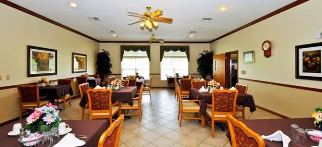 A dining room with several tables covered in dark tablecloths, each set with cups, napkins, and floral centerpieces. The room has tiled floors, ceiling fans with lights, framed artwork on the walls, and large windows with green valances letting in natural light.