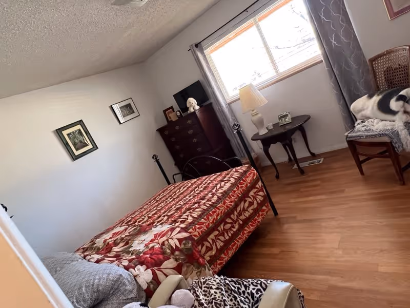 Small bedroom with a single metal-frame bed covered in a red floral quilt, dresser, side table and a chair by a window.