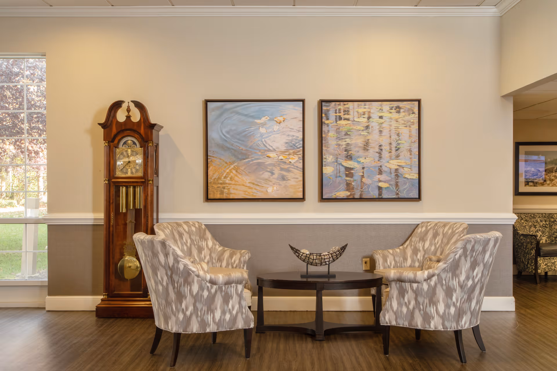 A cozy seating area in a senior living facility with two patterned armchairs facing each other across a dark wooden coffee table. Behind the chairs, there is a tall grandfather clock and two framed paintings depicting water with floating leaves. A large window on the left lets in natural light, and the walls are painted in neutral tones with white trim.