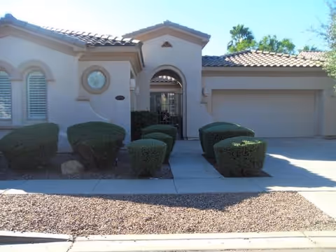 Exterior view of a single-story residential building with a tiled roof, arched entryway, and neatly trimmed bushes lining the walkway leading to the front door. The driveway and garage are visible on the right side.