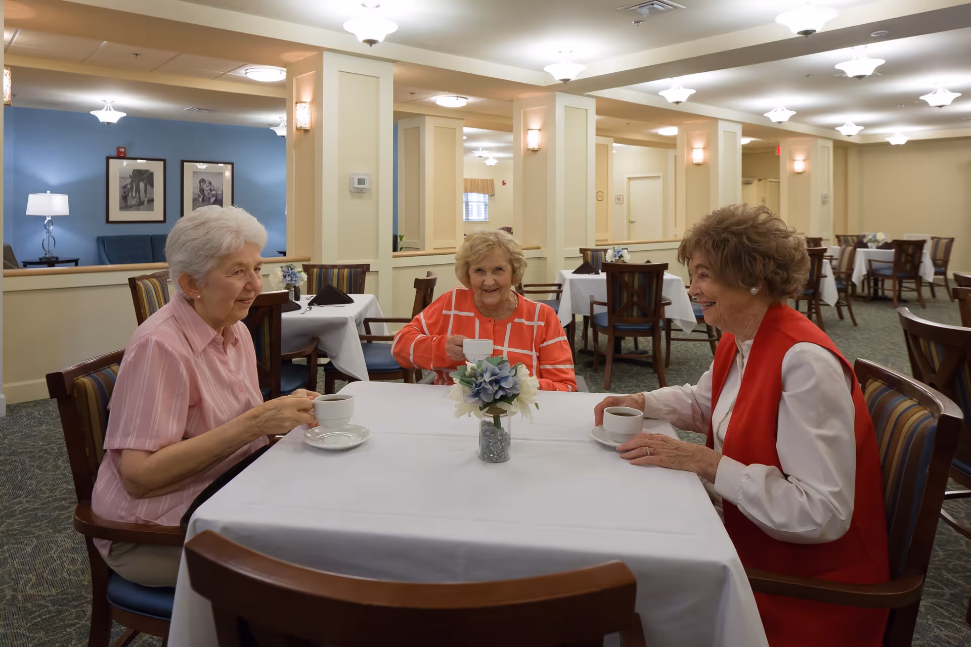 Three elderly women sitting around a table in a dining room, each holding a cup of coffee or tea. The table is covered with a white tablecloth and has a small floral centerpiece. The room is well-lit with ceiling lights and has multiple tables and chairs arranged neatly.