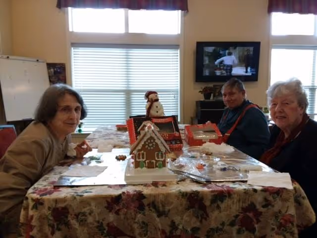 Three elderly individuals sitting around a table covered with a floral tablecloth, engaging in decorating gingerbread houses. There is a snowman decoration on the table, and a television is visible in the background with a person on the screen. The room has large windows with blinds and valances.