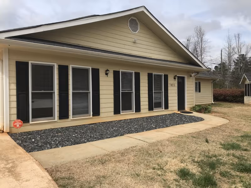Single-story beige house with black shutters and a paved walkway leading to the front door and windows, set in a grassy yard.