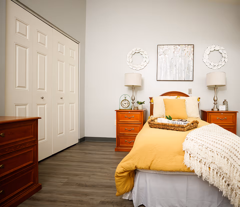 A cozy bedroom with a single bed covered in a yellow comforter and a white knitted throw blanket. The bed has a yellow pillow and a wicker tray with books and glasses on it. On either side of the bed are wooden nightstands with matching lamps, a clock, and small decorative items. Above the bed hangs a framed artwork of trees, flanked by two white wreath decorations. To the left, there is a wooden dresser and a set of white closet doors. The room has light-colored walls and wood flooring.