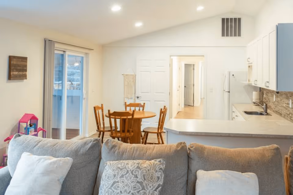 Open-plan living area with a couch in the foreground, a wooden dining table and chairs in the middle, and a white kitchen with cabinets and a countertop in the background.