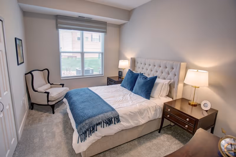 A cozy bedroom with a large bed featuring a tufted headboard, white bedding, and blue accent pillows and throw. There are two wooden nightstands on either side of the bed, each with a lamp. A cushioned armchair is placed near a window with blinds, and the room has light gray walls and carpeted flooring.