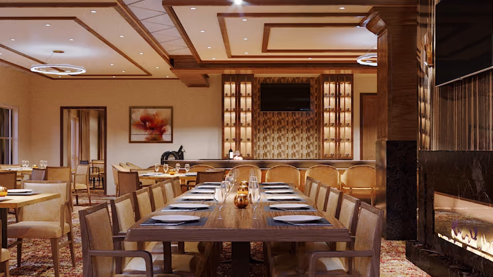 A spacious dining room with a long wooden table set with plates, glasses, and small decorative pumpkins. The room features beige upholstered chairs, a patterned carpet, a fireplace on the right, and a wall-mounted TV above a wooden cabinet with glass shelves. Warm lighting and a ceiling with wooden beams add to the cozy ambiance.