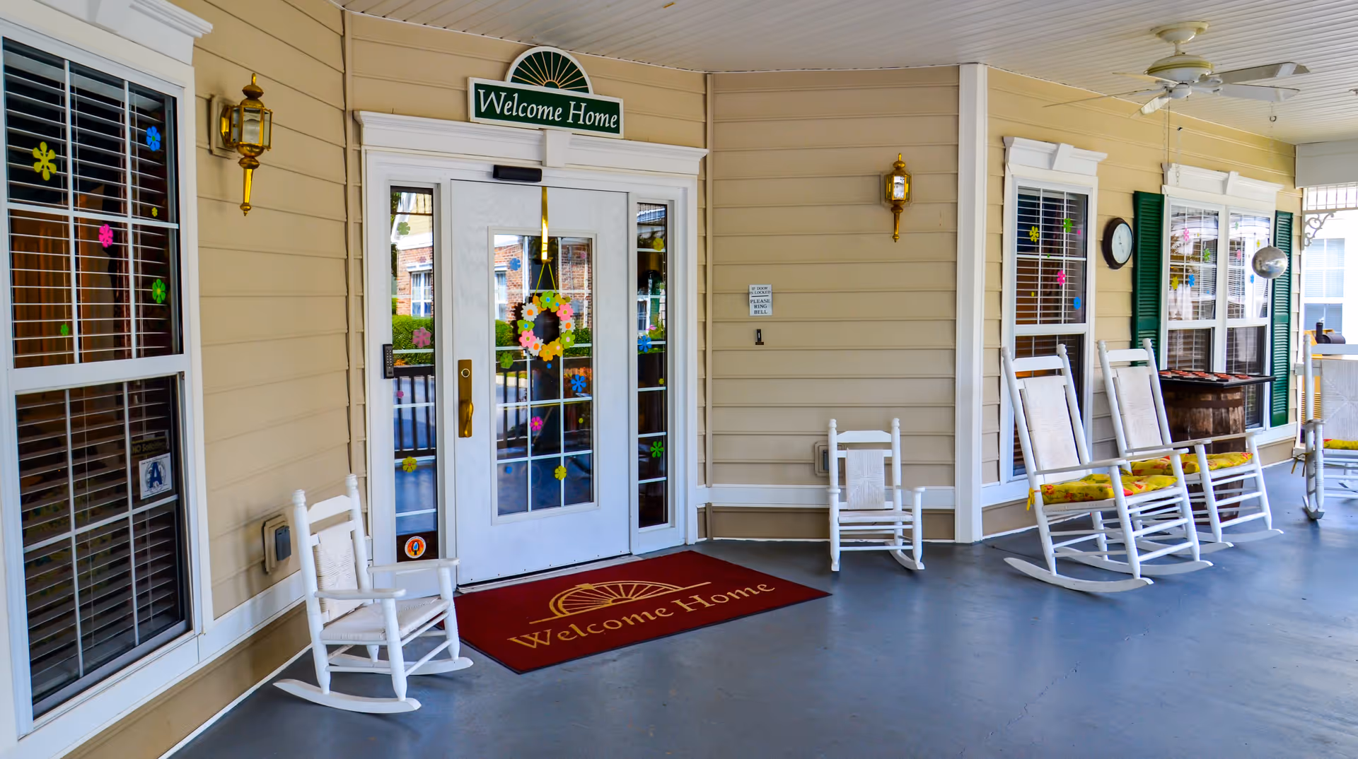 Covered porch entrance of a facility with beige siding, white framed windows decorated with colorful flower stickers, and a white double door with glass panels. Above the door is a green sign that reads 'Welcome Home' and a matching red welcome mat on the floor. There are several white rocking chairs with cushions arranged along the porch.