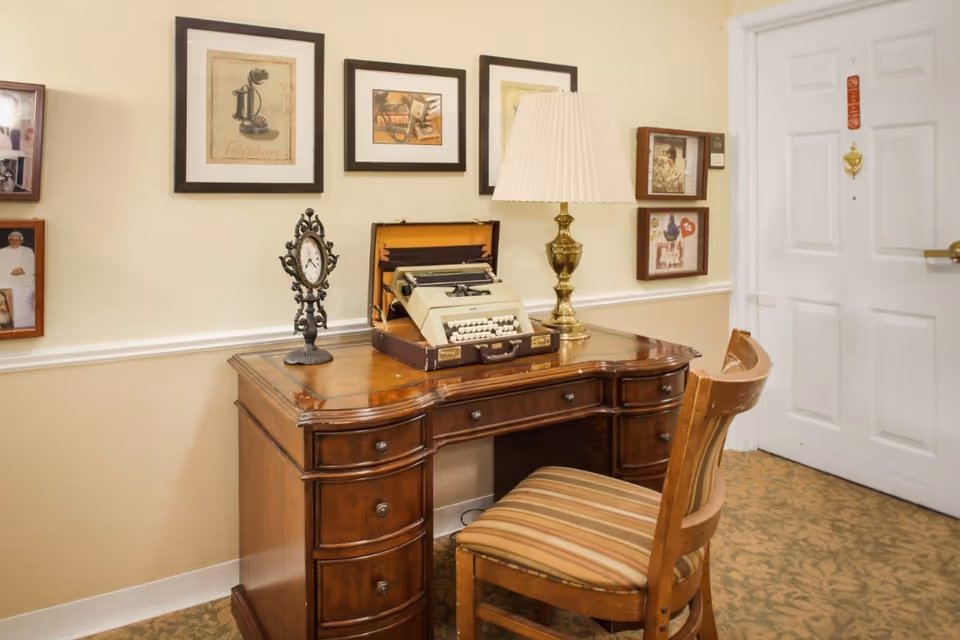 A vintage wooden desk with multiple drawers, featuring an old typewriter, a decorative clock, and a brass table lamp with a pleated shade. A wooden chair with a striped cushion is placed in front of the desk. The wall behind the desk is decorated with framed pictures and artwork, and a white door is visible to the right.