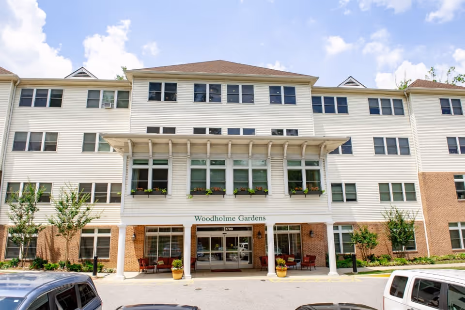 Front exterior view of Woodholme Gardens, a multi-story senior living facility with white siding and brick accents. The entrance features a covered porch supported by white columns, with potted plants and outdoor seating on either side. Several cars are parked in front of the building under a partly cloudy sky.