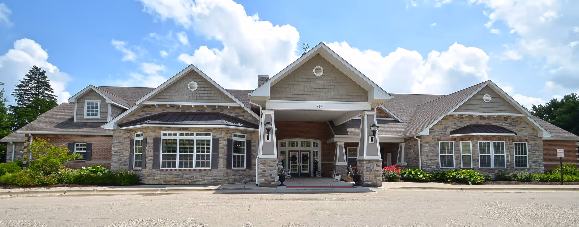 Front exterior view of a single-story senior living facility building with stone and brick facade, large windows, a covered entrance with pillars, and a clear blue sky with clouds above.