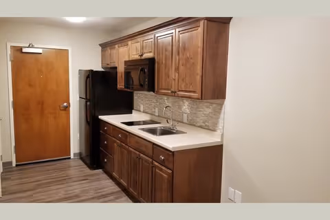 Small kitchen area with wooden cabinets, a countertop with sink, black refrigerator and microwave, and an entry door at the end of the corridor.