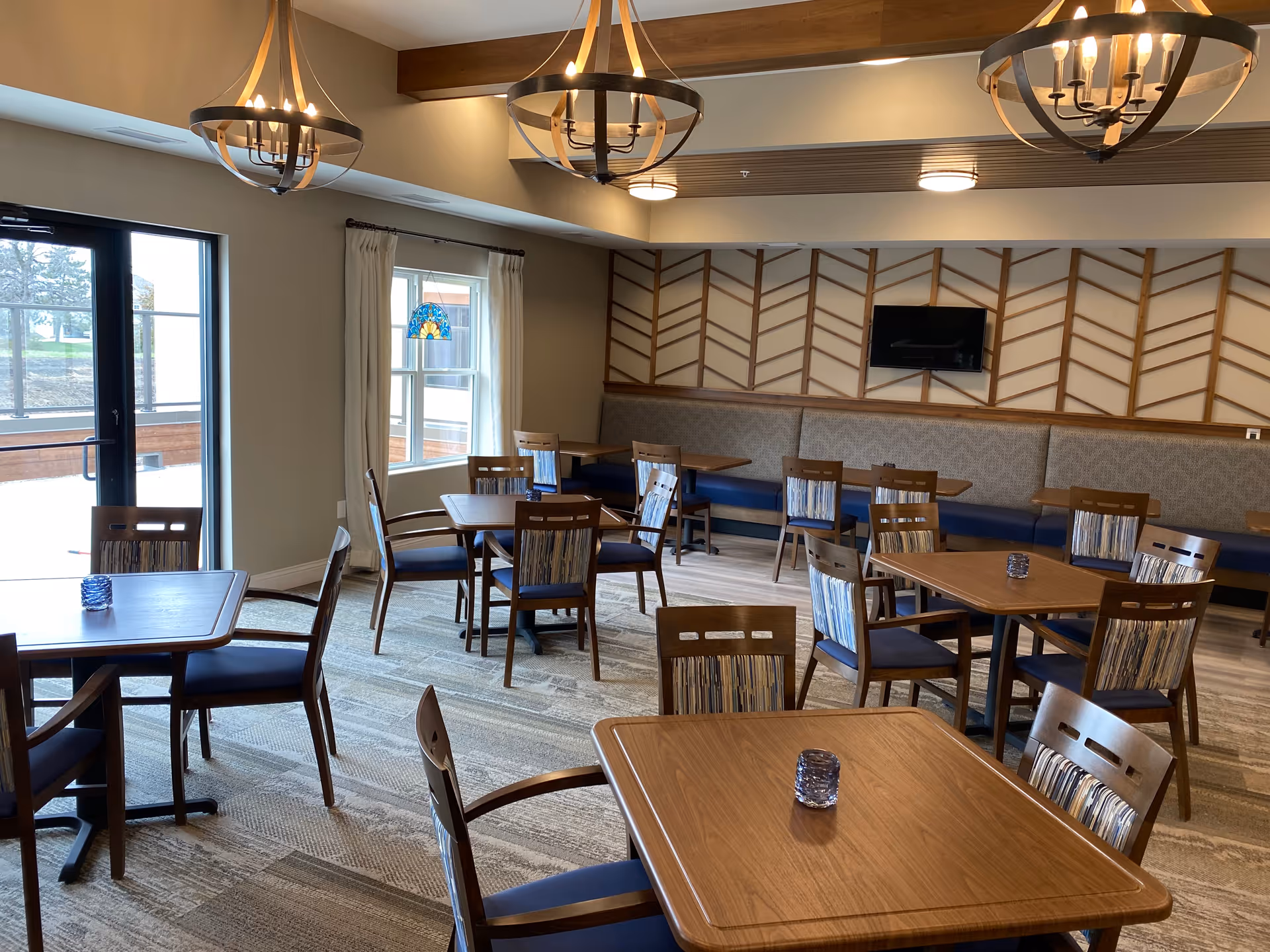 A well-lit dining room with multiple wooden tables and chairs featuring blue cushions. The room has large windows with white curtains, modern chandeliers hanging from the ceiling, and a wall-mounted TV on a decorative wooden panel wall. The floor is carpeted with a patterned design.