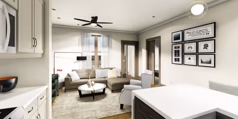 A modern living room with a sectional sofa, an armchair, a round coffee table, and a ceiling fan. The room has light-colored walls with framed pictures and quotes on one wall. There is a window with sheer curtains and a door leading outside. The foreground shows part of a kitchen island with a white countertop and cabinetry.