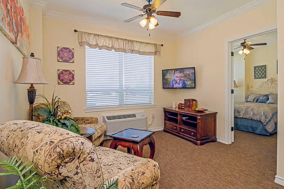 A cozy living room with floral patterned sofa and armchair, a wooden coffee table, a floor lamp, and a TV mounted on the wall. There is a window with blinds and a valance, and a doorway leading to a bedroom with a bed and ceiling fan visible.