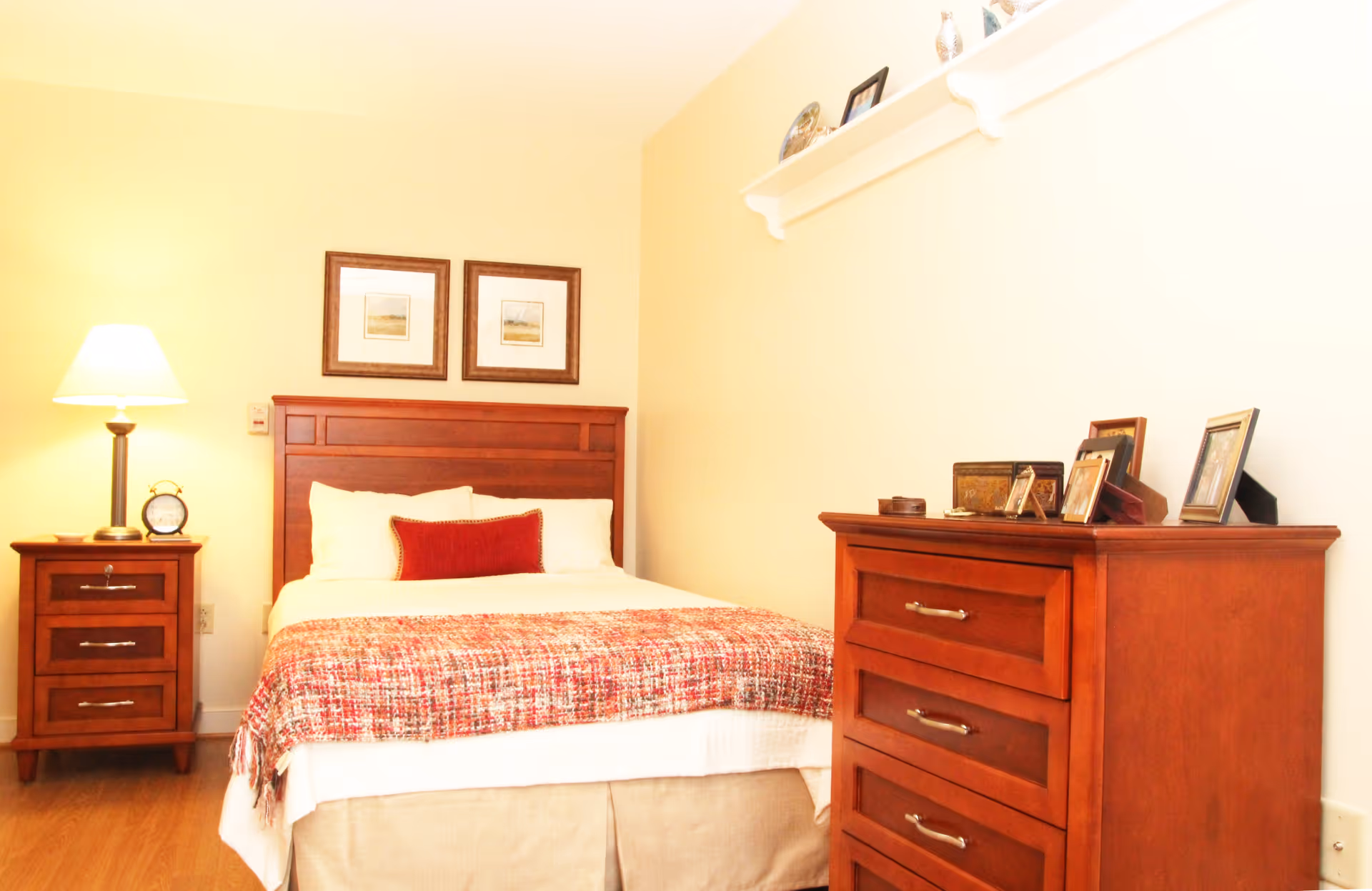 A cozy bedroom with a wooden bed frame, a bed made with white sheets, a red decorative pillow, and a red patterned throw blanket. There is a wooden nightstand with a lamp and an alarm clock on the left side of the bed. On the right side of the room, there is a wooden dresser with several framed photos and decorative items on top. Two framed pictures hang on the wall above the bed, and a small white shelf with decorative items is mounted on the adjacent wall.