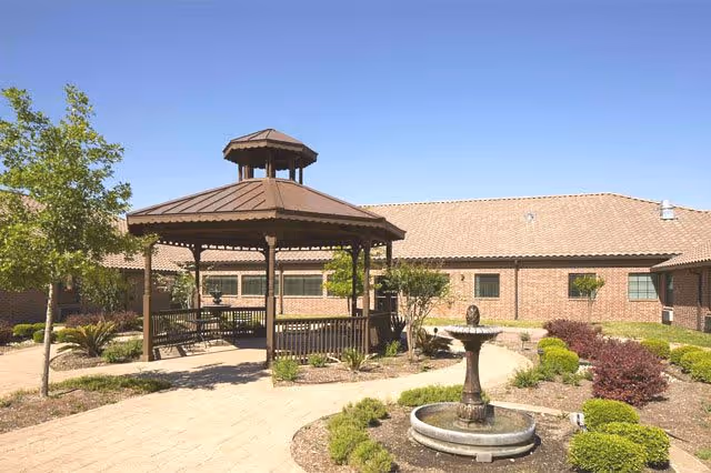 Outdoor courtyard area with a wooden gazebo, surrounded by landscaped plants and bushes, a stone pathway, and a water fountain in front. The background shows a single-story brick building under a clear blue sky.