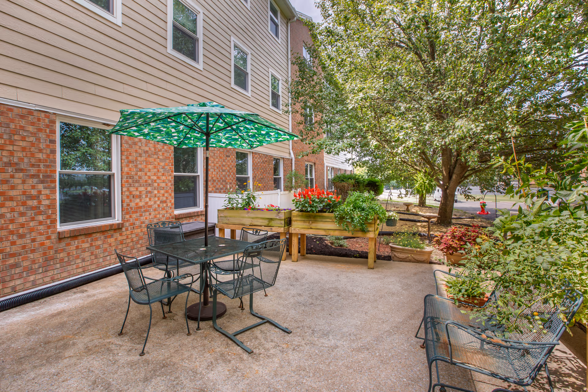 Outdoor patio area with a metal table and four chairs under a green patterned umbrella. Raised garden beds with colorful flowers and plants are nearby, along with additional potted plants and a large tree providing shade. The patio is adjacent to a brick and siding building with several windows.