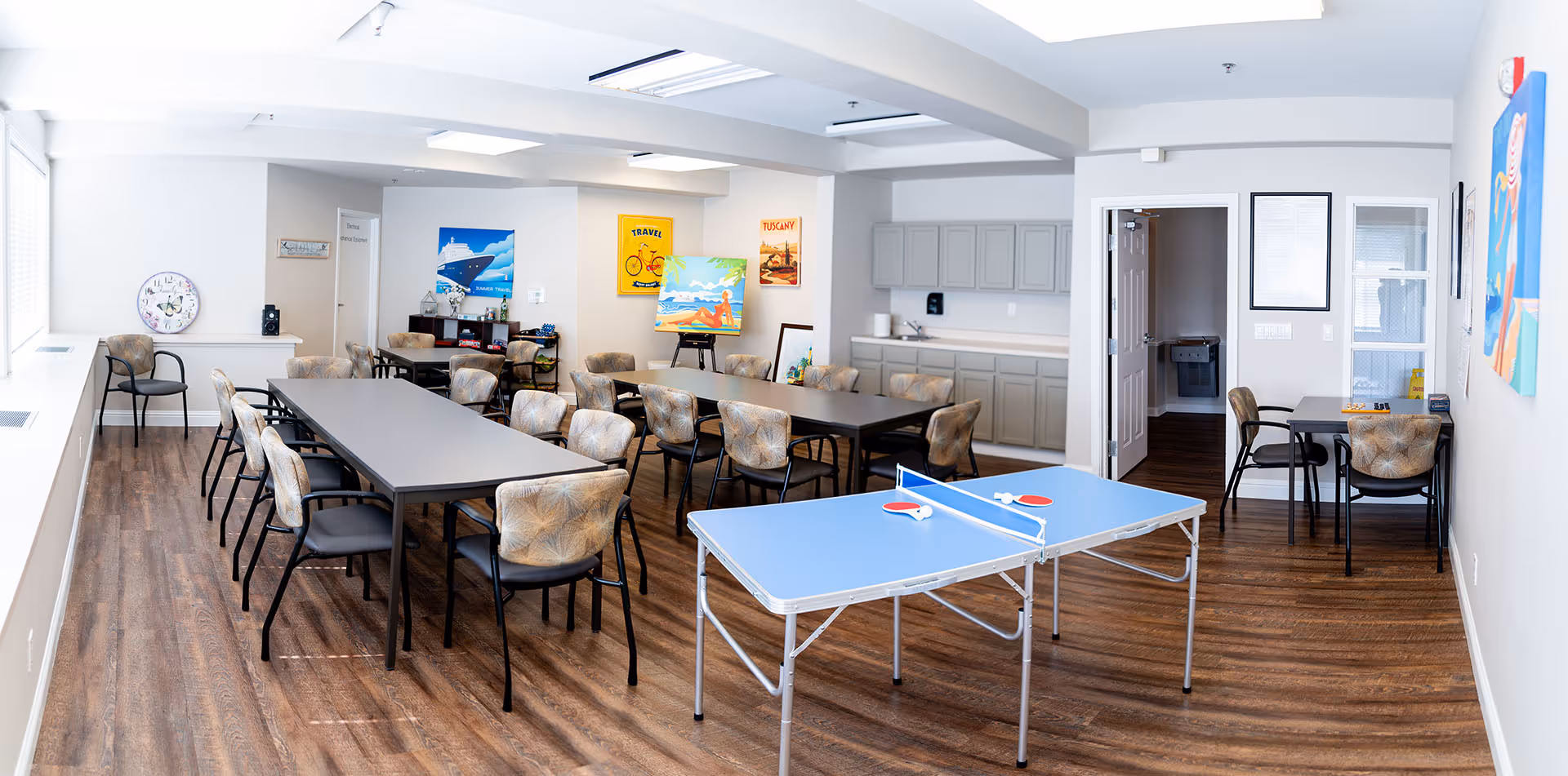 Bright multipurpose senior activity room with long tables and chairs, a kitchenette, artwork on the walls, and a ping-pong table in the foreground.
