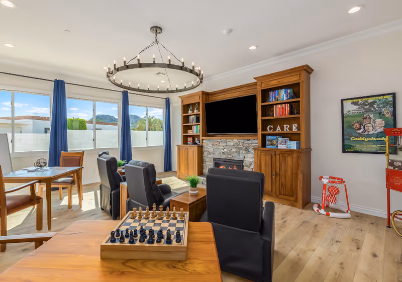 A bright and cozy common living area with large windows and blue curtains letting in natural light. The room features a wooden entertainment center with a TV above a stone fireplace, bookshelves with books and decorative items, and the word CARE displayed on one shelf. There are several comfortable black armchairs arranged around a wooden coffee table with a small plant. A chessboard is set up on a nearby table. The room also includes a small basketball hoop and a popcorn machine, with a framed Caddyshack movie poster on the wall. The flooring is light wood, and a modern circular chandelier hangs from the ceiling.