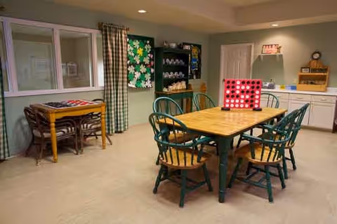 A cozy activity room with a wooden table and six green chairs in the center. On the table is a large Connect Four game. In the background, there is a small table with chairs, a black shelving unit with various items, a bulletin board with decorations, and a countertop with cabinets and a clock above it. The room has light-colored walls and flooring, with green and white checkered curtains on the window.