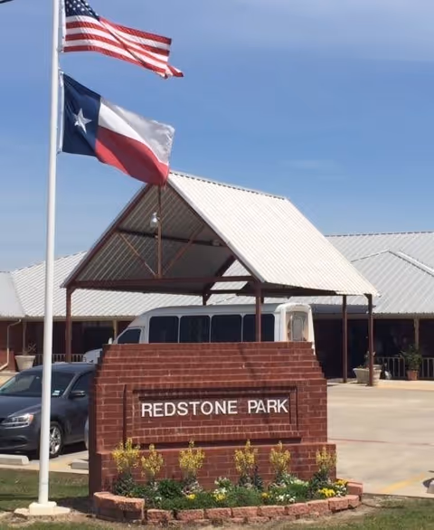 Outdoor view of the entrance area of Redstone Park facility with a brick sign displaying the name 'REDSTONE PARK'. Two flagpoles are visible, one with the American flag and the other with the Texas state flag. A covered structure with a metal roof is behind the sign, with a parked vehicle and part of the building visible in the background under a clear blue sky.