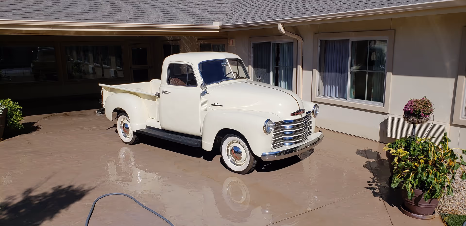 A vintage white pickup truck parked on a concrete driveway outside a building with beige walls and windows. There are potted plants on the right side near the building, and a garden hose is visible on the ground in the foreground.