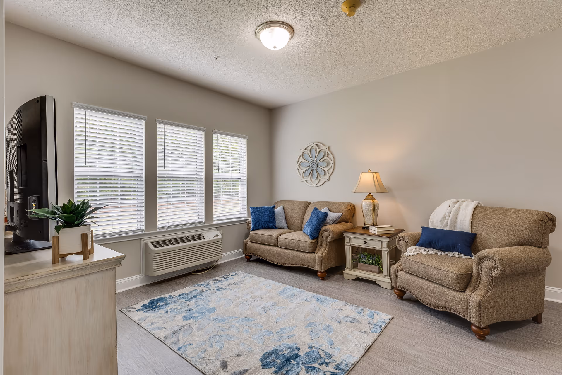 A cozy living room with two beige upholstered armchairs and a loveseat, each adorned with blue and gray throw pillows. A small side table with a lamp and books sits between the loveseat and one armchair. A decorative wall piece hangs above the loveseat. The room has three windows with white blinds, a light-colored area rug with blue floral patterns on the floor, and a TV on a light-colored stand with a small potted plant. The walls are painted light gray and the floor is a light wood tone.
