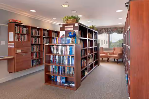 Interior view of a library or reading room with wooden bookshelves filled with books. There are two upholstered chairs and a small table near a window with a valance curtain. The room is well-lit with ceiling lights and has a carpeted floor.