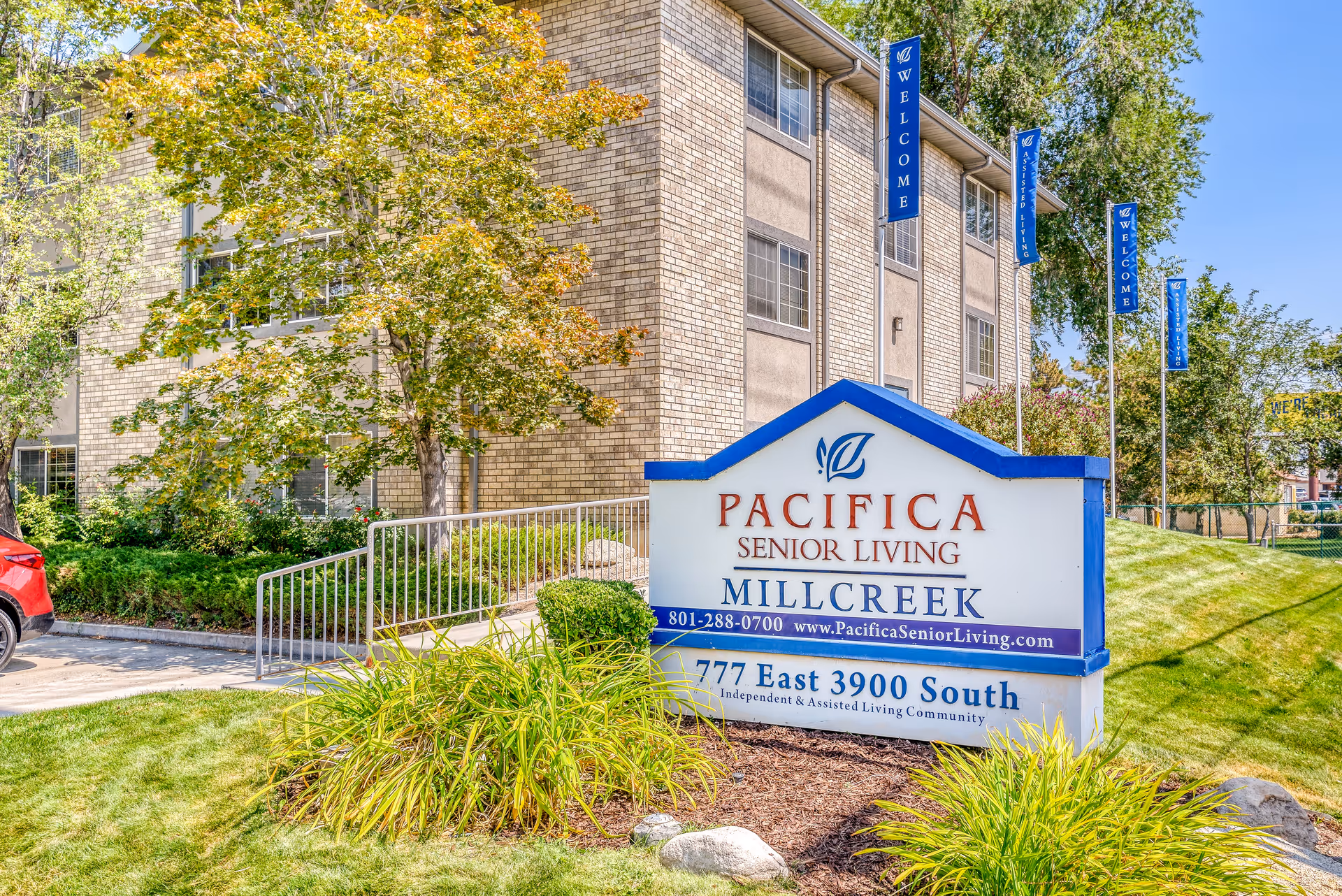 Exterior view of Pacifica Senior Living Millcreek building with a large sign in front displaying the facility name, contact number, website, and address. The building is surrounded by green grass, trees, and shrubs under a clear blue sky.