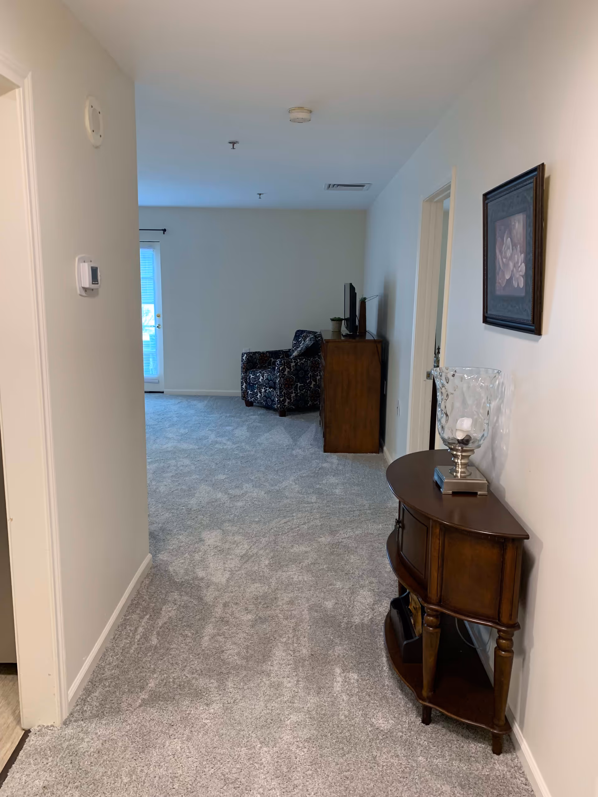 View down a carpeted hallway leading to a living area with a patterned armchair and a wooden cabinet with a TV on top. On the right side of the hallway, there is a small wooden console table with a glass decorative piece and a framed floral picture hanging above it. The walls are painted white and there is a thermostat on the left wall.