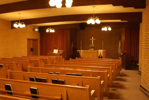 A chapel-like interior with rows of wooden pews facing an altar topped by a cross and lit by hanging fixtures.