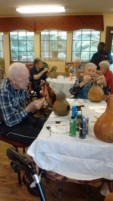 A group of elderly individuals seated around tables in a well-lit room, engaging in an arts and crafts activity involving large gourds. Various art supplies such as paint bottles, brushes, and palettes are on the tables. Large windows with red valances allow natural light into the room.