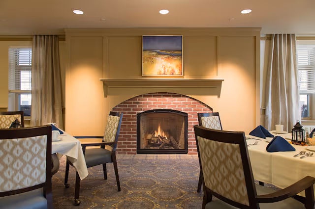 Cozy dining room with tables set for service and chairs facing a brick fireplace beneath a framed painting.