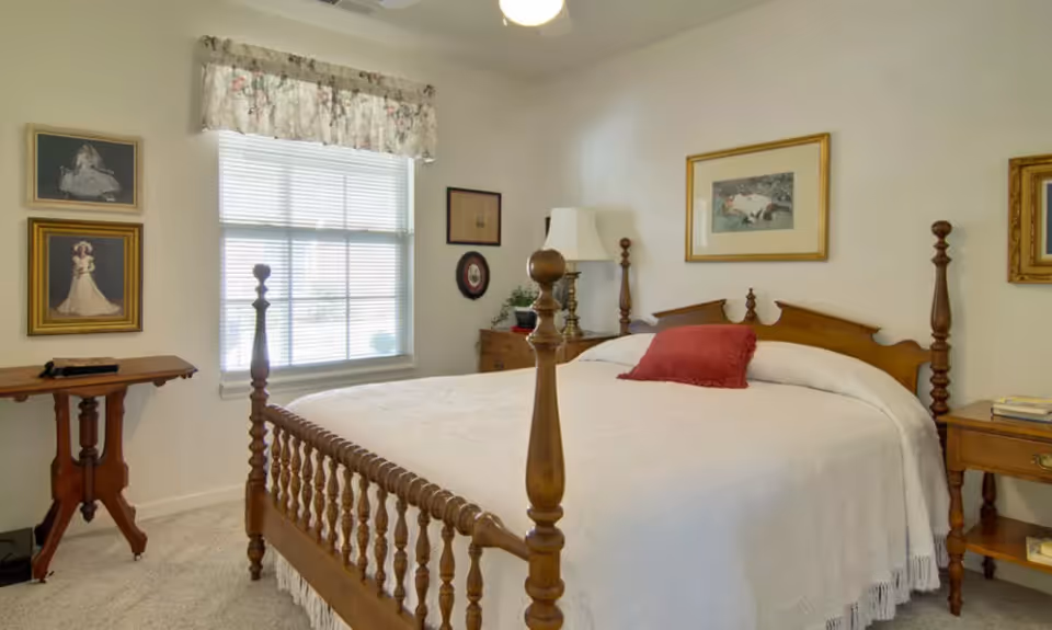 A bright bedroom with a wooden four‑poster bed dressed in a white bedspread, bedside tables, framed artwork, and a window with blinds.