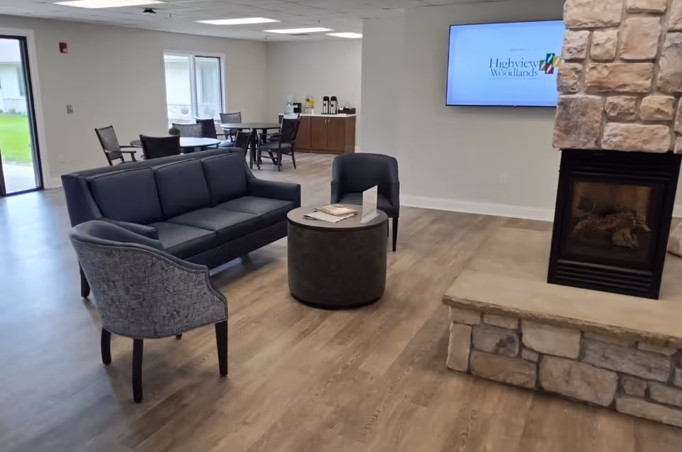 A modern living room area with a dark gray sofa, two armchairs, and a round coffee table in the center. The room has wood flooring and a stone fireplace on the right. In the background, there are tables and chairs near windows and a cabinet with coffee and beverage supplies. A wall-mounted TV displays the text 'Welcome to Highview Woodlands'.