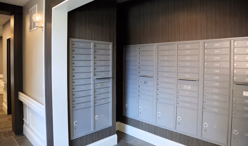 Wall of numbered metal mailboxes mounted in an interior hallway with tiled floor and decorative trim.