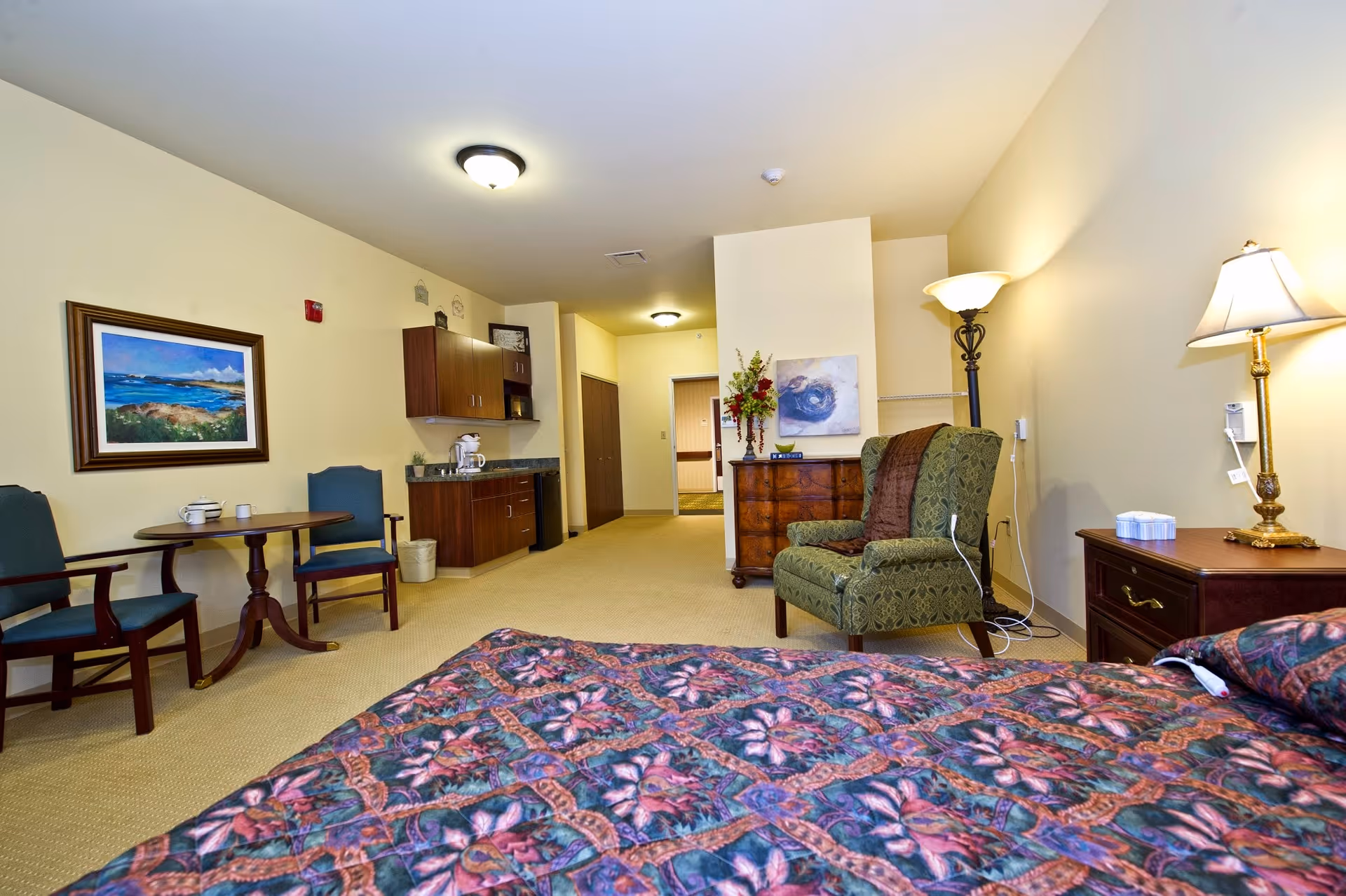 A spacious senior living bedroom featuring a bed with a colorful floral bedspread in the foreground. The room includes a green upholstered armchair with a brown throw, a wooden dresser with a vase of flowers and a painting above it, a small round table with two chairs, a kitchenette area with cabinets, a coffee maker, and a trash bin. The walls are painted light yellow, and the room is well-lit with ceiling and floor lamps.