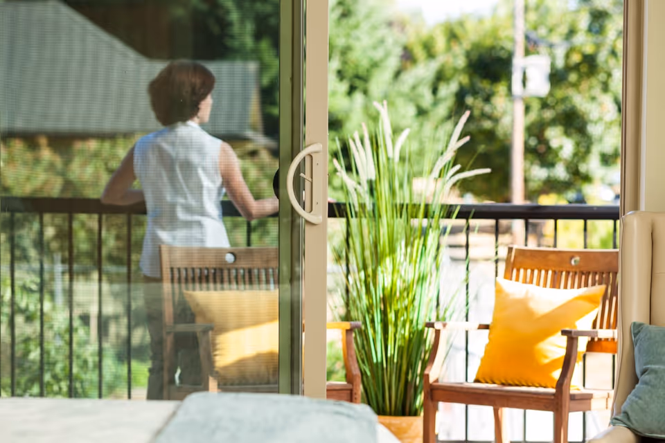 View through a sliding glass door to a sunlit balcony with wooden chairs, yellow cushions, a tall potted plant, and a person standing outside.