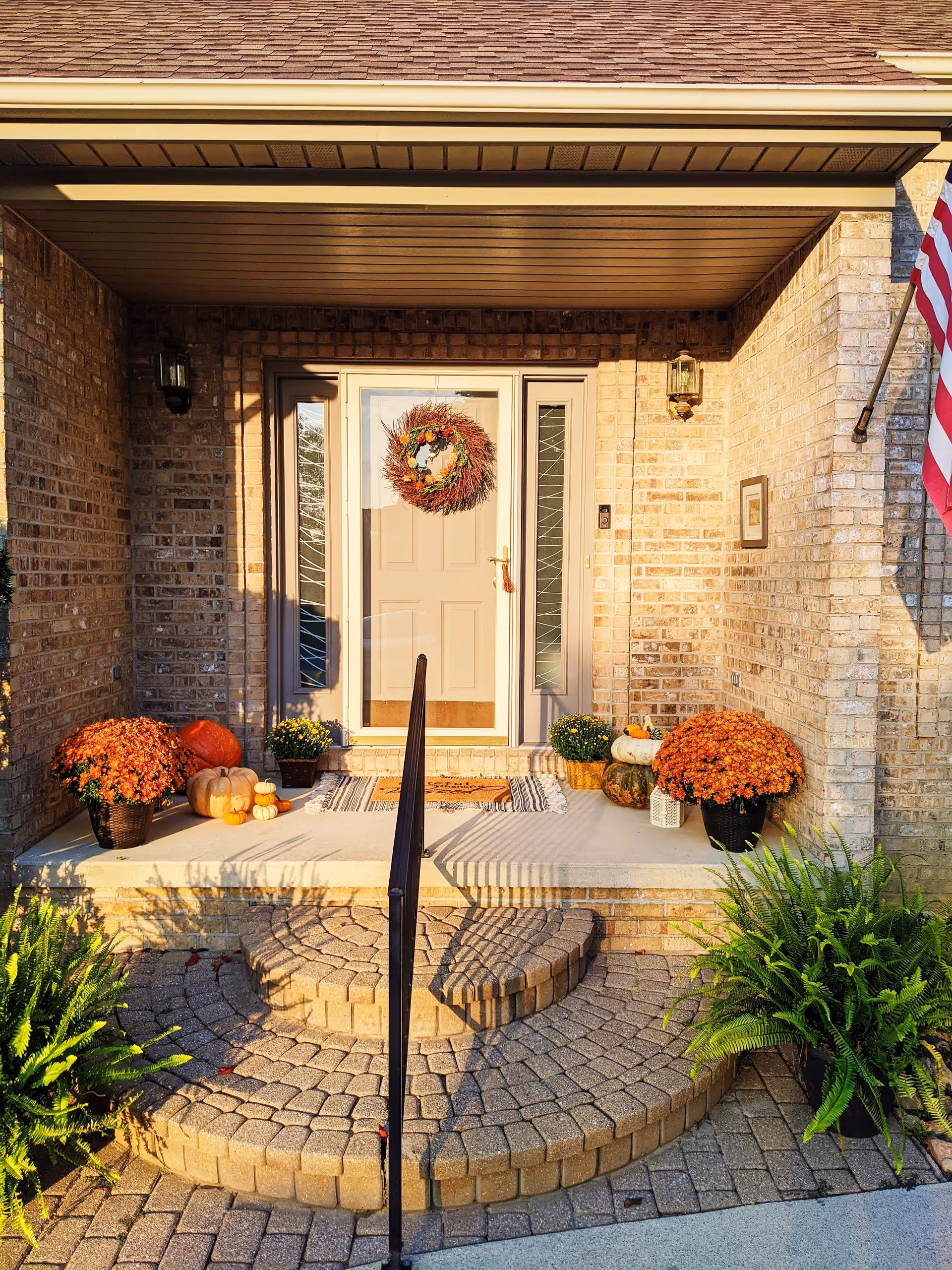 Front entrance of a brick building with a beige door decorated with a fall-themed wreath. The porch has potted orange chrysanthemums, pumpkins, and a welcome mat. There are stone steps leading up to the porch with a black handrail in the center. An American flag is visible on the right side.