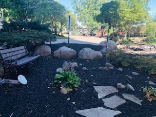 Outdoor garden area with a wooden bench on the left, a stone pathway leading through mulch and small plants, surrounded by trees and greenery under a clear sky.