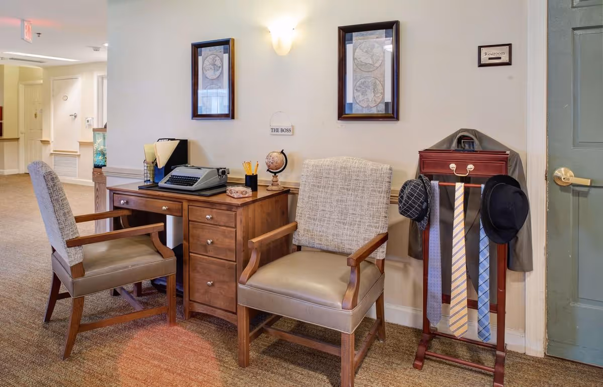 A cozy office corner in a senior living facility with a wooden desk holding a vintage typewriter, a small globe, pencils, and file folders. Two cushioned wooden chairs are placed in front of the desk. On the wall above the desk are two framed maps and a small sign that reads 'THE BOSS'. To the right, a wooden valet stand holds a jacket, three neckties, and two hats. A door with a sign labeled 'Restroom' is visible on the right side.