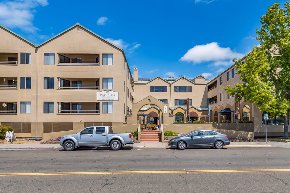 Exterior view of a multi-story senior living facility with a central entrance and parked cars.