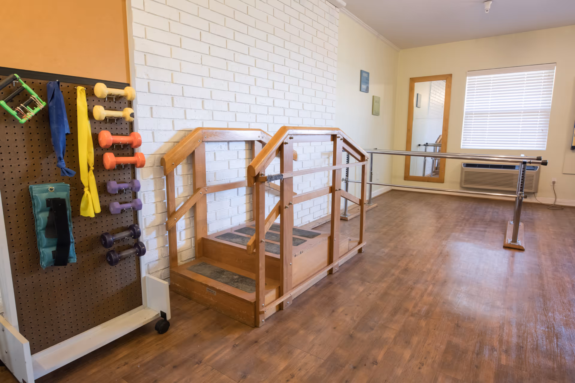 A senior living facility exercise room with wooden stairs and handrails for physical therapy, a ballet barre along the wall, a large mirror, a window with blinds, and a pegboard holding various exercise equipment including dumbbells and resistance bands.