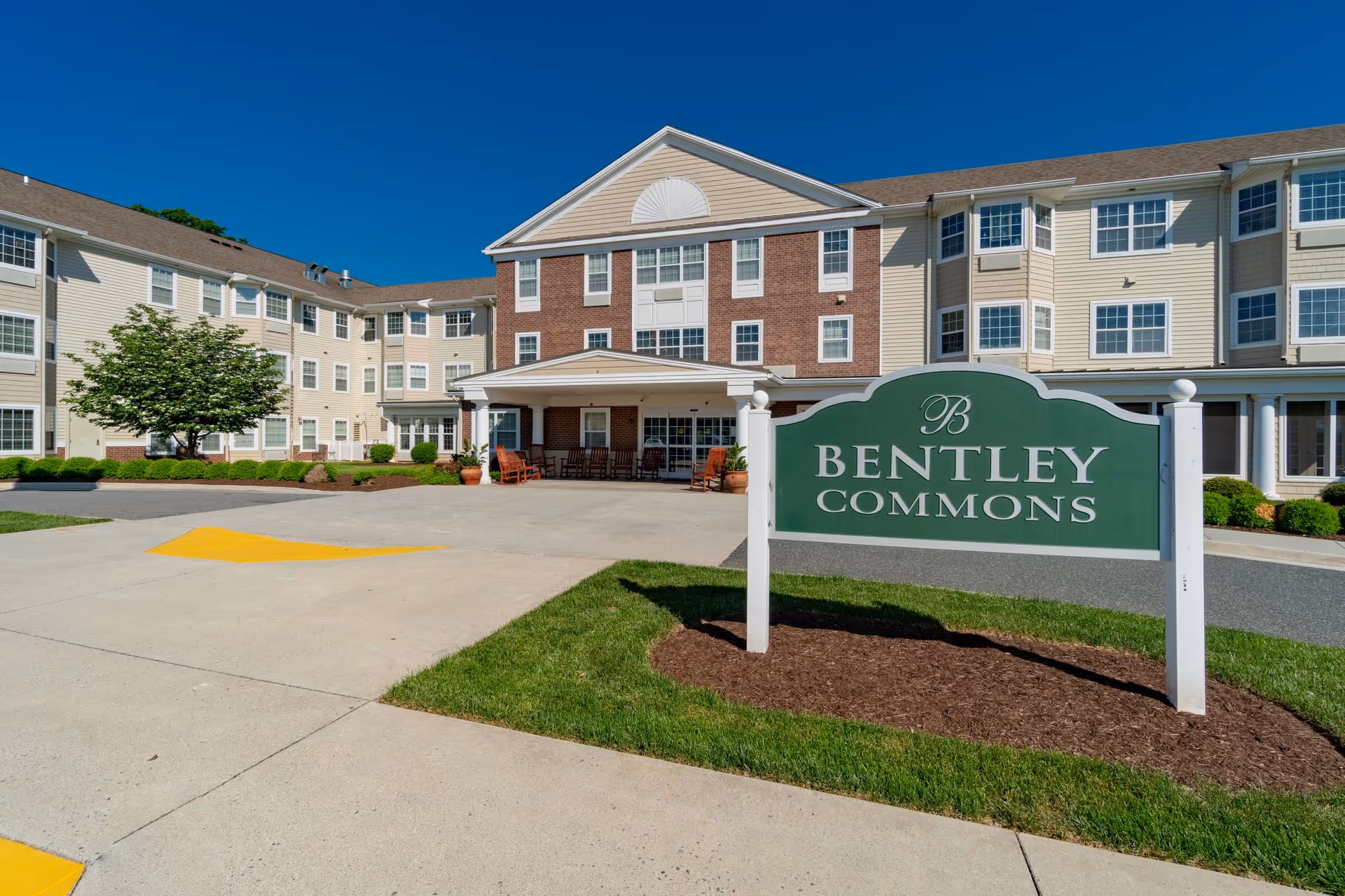 Exterior view of Bentley Commons at Lynchburg, a multi-story senior living facility with beige and brick facade, large windows, a covered entrance with seating, and a green sign with white text displaying the facility name in front on a landscaped lawn.