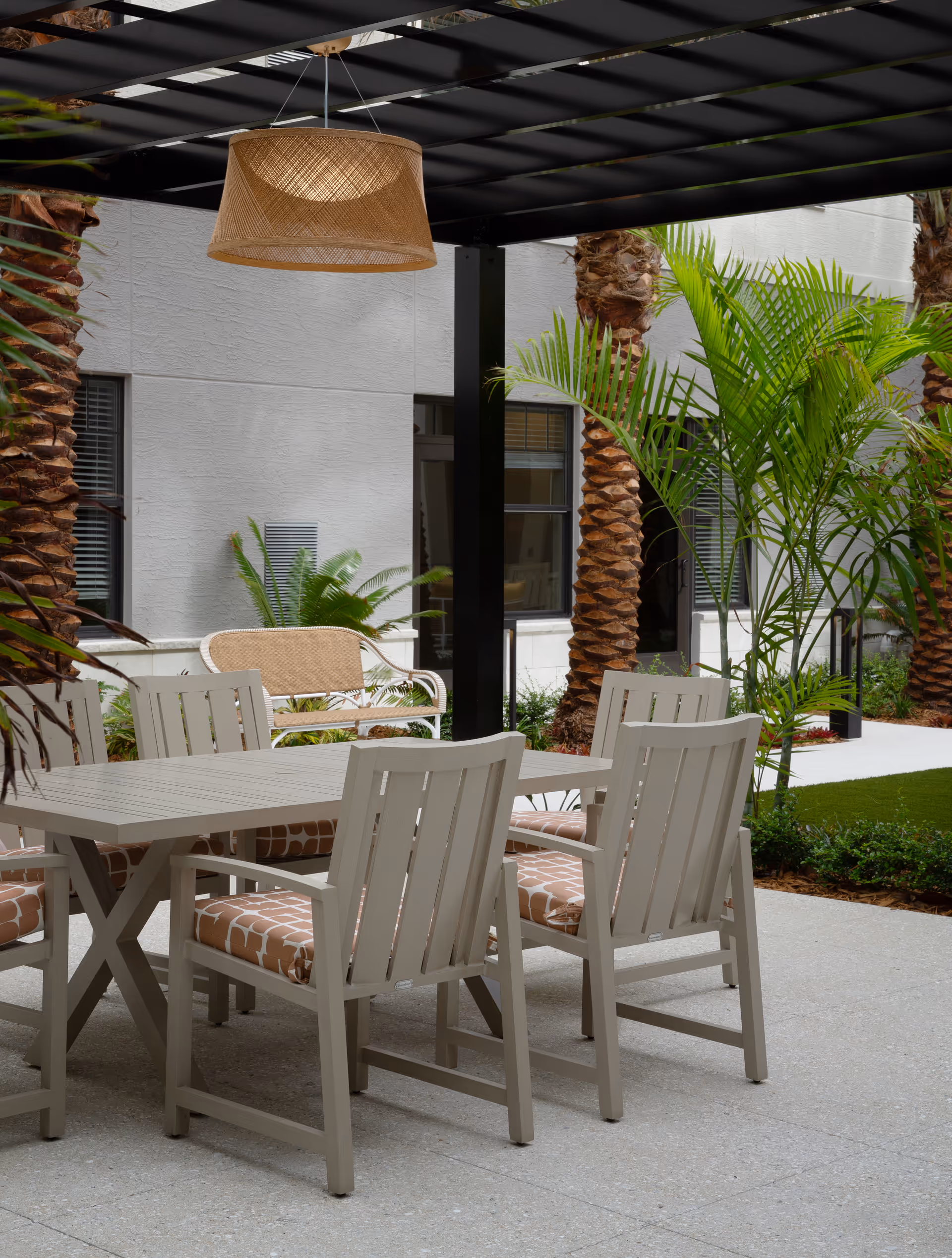 Outdoor patio area with a rectangular table surrounded by six cushioned chairs under a black pergola. There are palm trees and green plants around the patio, with a building wall and windows in the background.